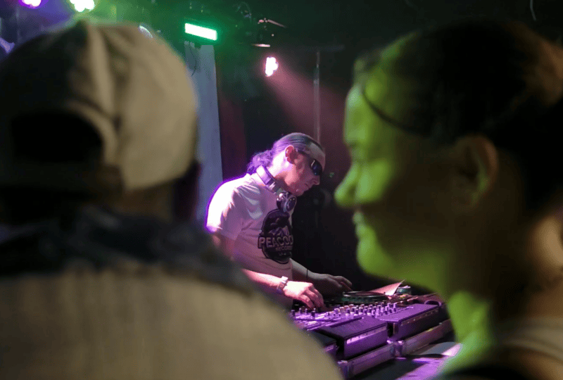 An image from behind two people dancing, focused on a DJ who is leaning over his gear on stage and performing